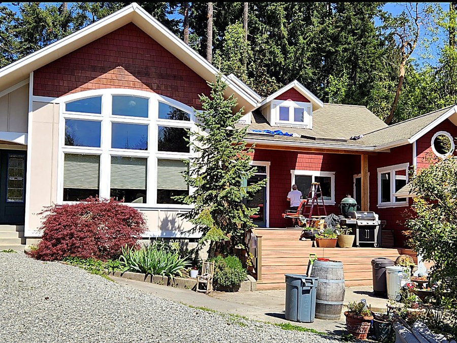 Two-story home clad in red cedar shingles with white window trim, a painter on a ladder cutting in trim beside the back deck.
