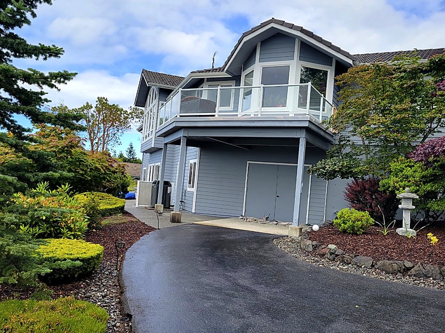 Two-story grey-blue home with crisp white trim and a glass balcony rail above the garage, seen from the landscaped driveway.